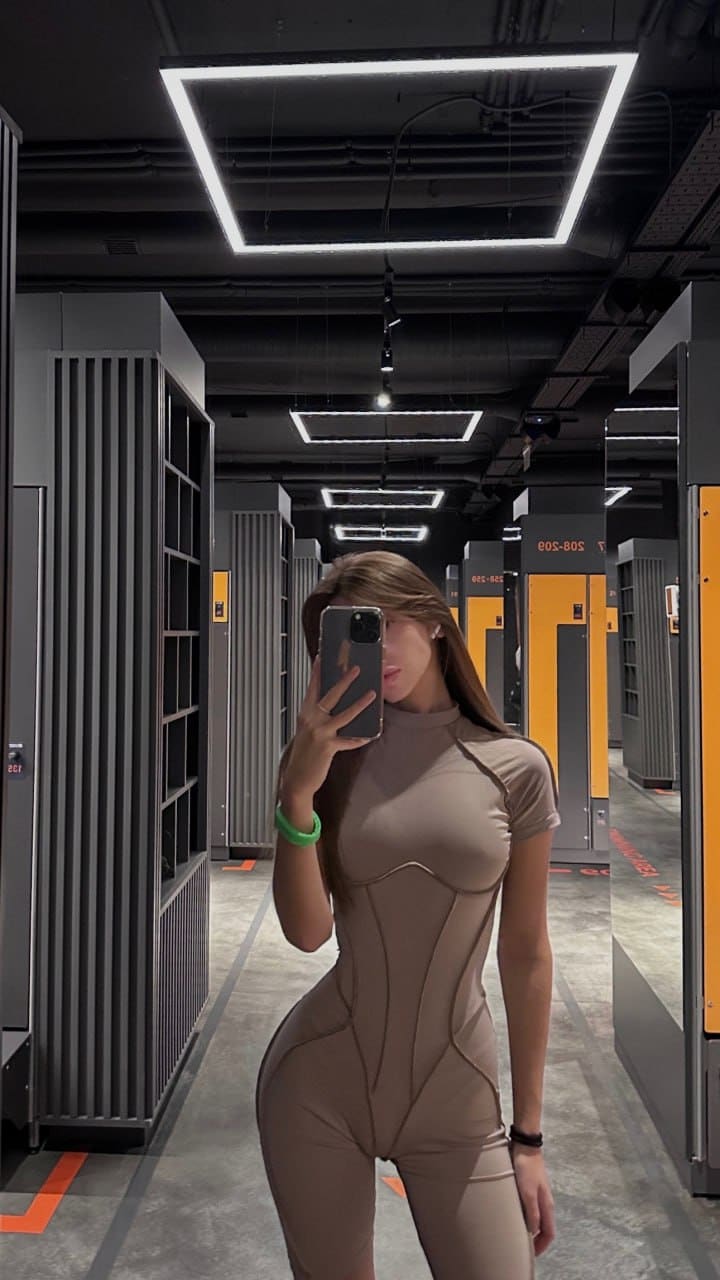 Женщина в бежевом боди с высоким воротом фотографирует себя в зеркало в проходе склада.