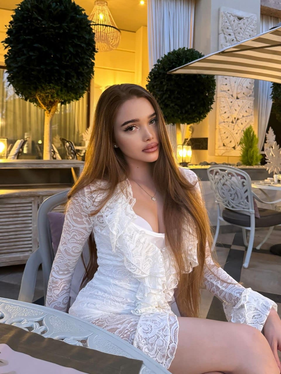 Арабелла в белом кружевном платье сидит за столом на террасе с топиариями и полосатым зонтом на фоне.