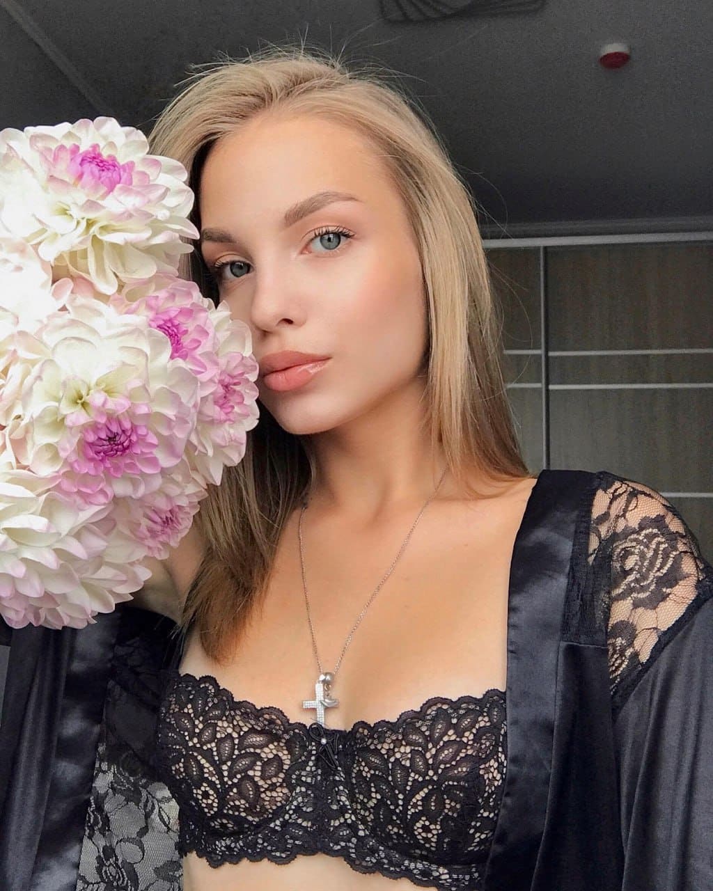 Карина в чёрном кружевном белье и халате держит букет белых и розовых цветов на фоне тёмной стены с сеткой.