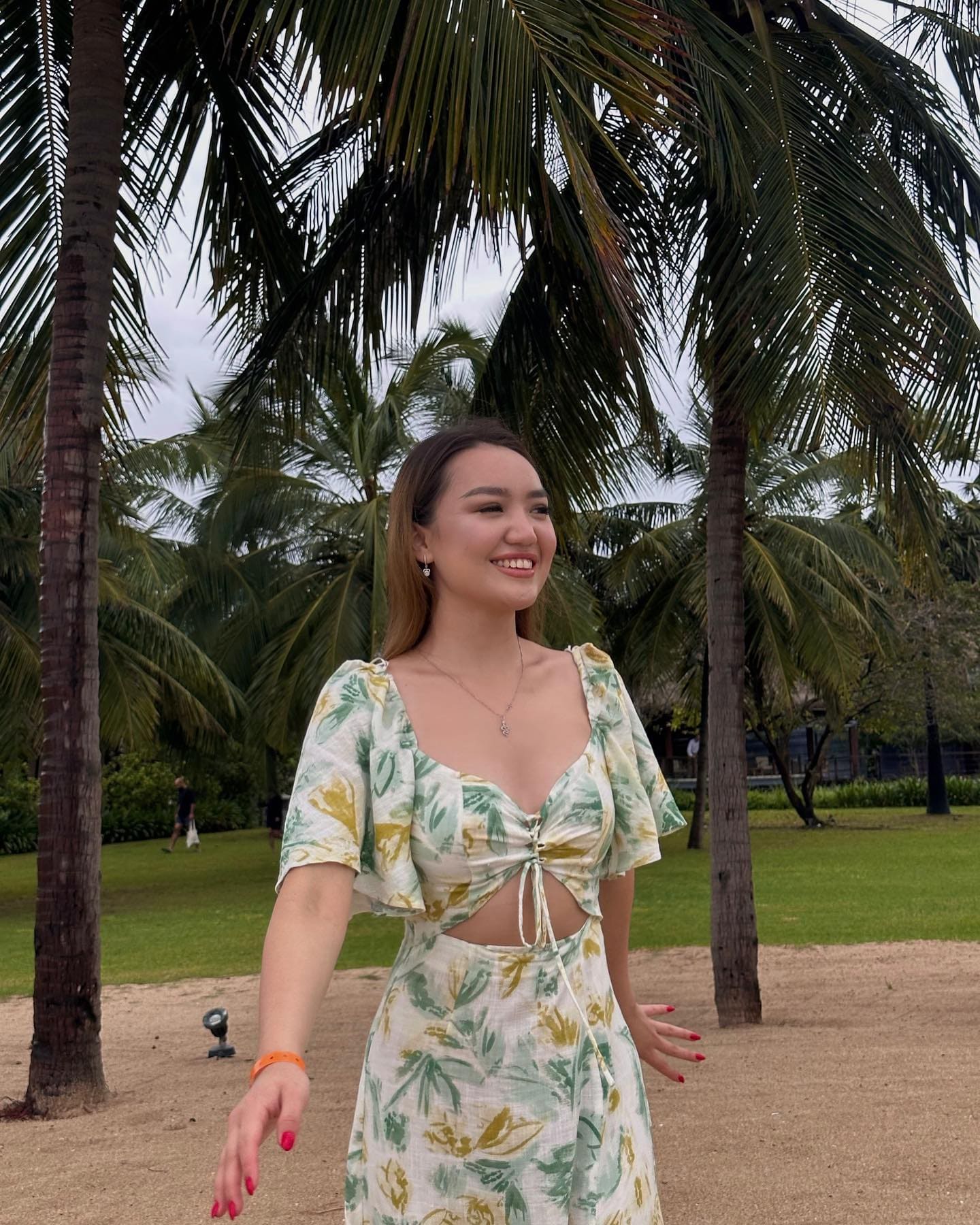 Женщина в цветочном костюме с вырезом на талии стоит на песчаной тропе среди пальм.